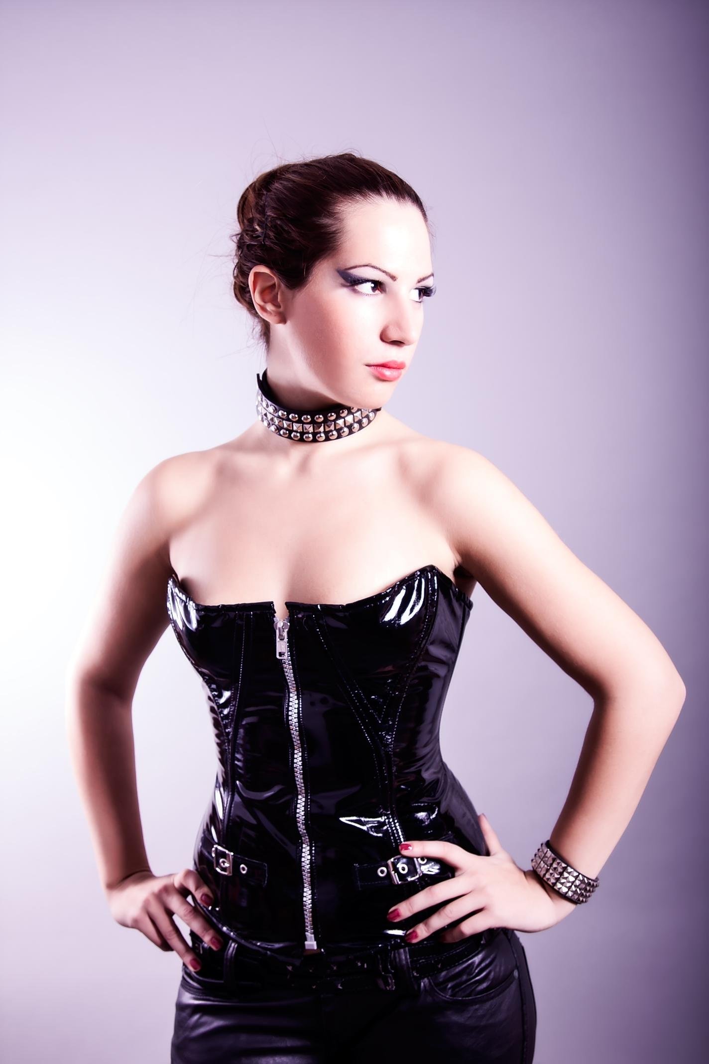 Woman in a shiny black PVC corset and leather pants, hands on hips, wearing studded choker and bracelet, looking defiantly to the side.