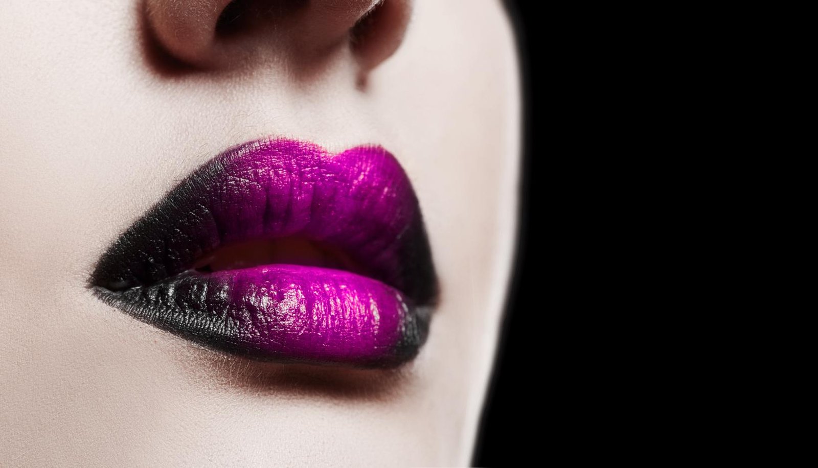 Close-up of glossy ombré lips with deep black fading into vibrant purple on porcelain skin, against a black background.