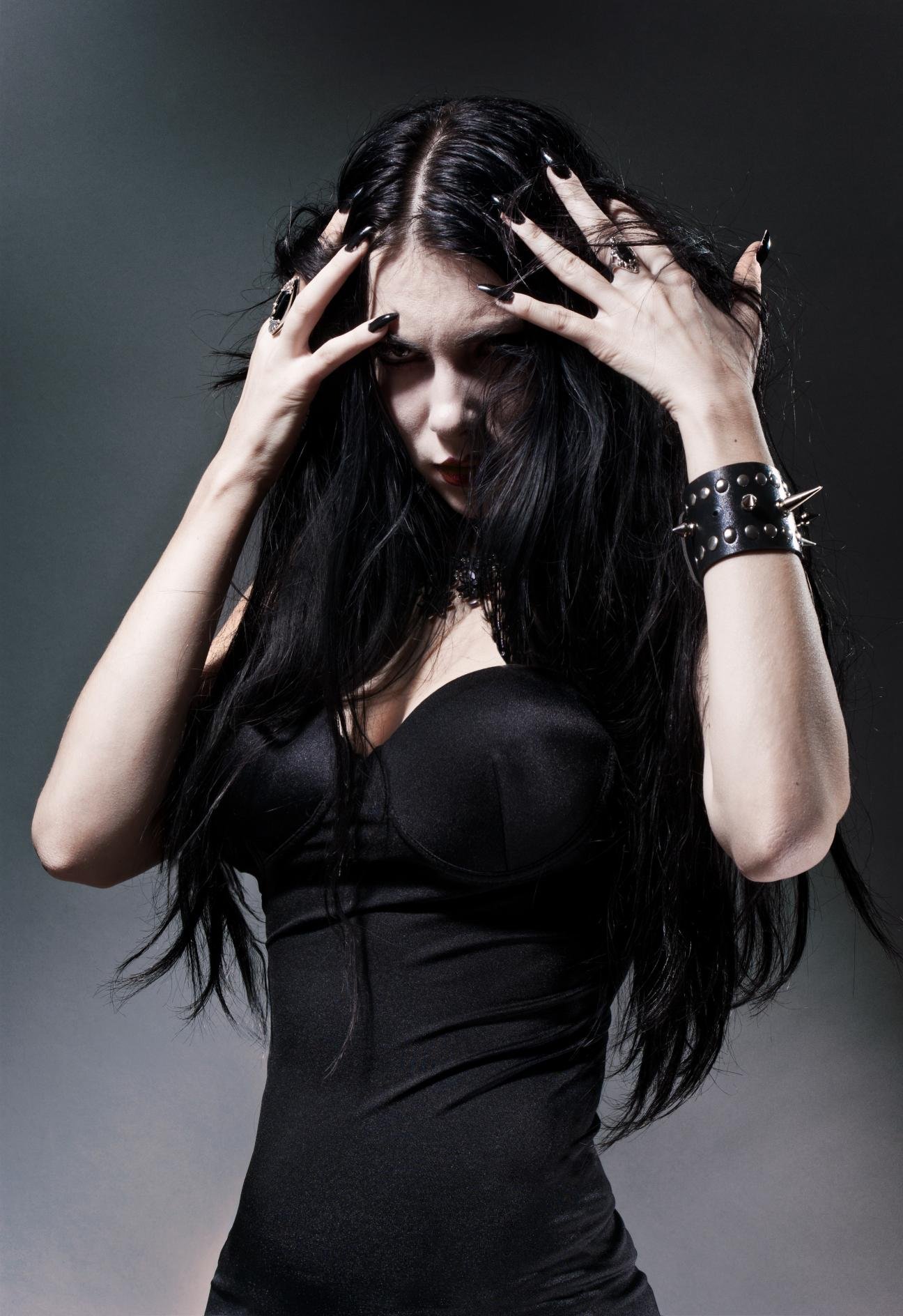 Gothic woman in a tight black corset clutching her head, long black hair and dark eye makeup, wearing spiked leather accessories.