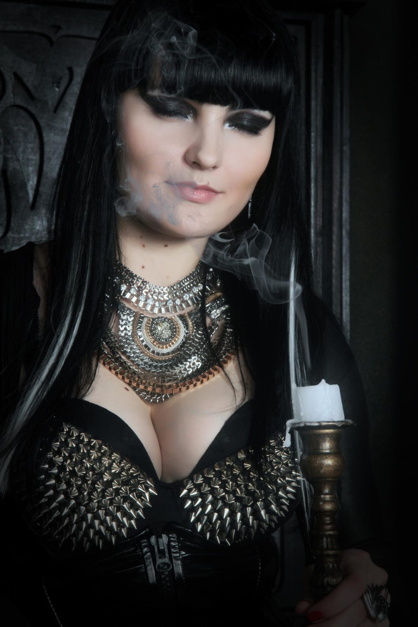 Gothic woman in studded black corset smoking beside candle, dramatic metal necklace.