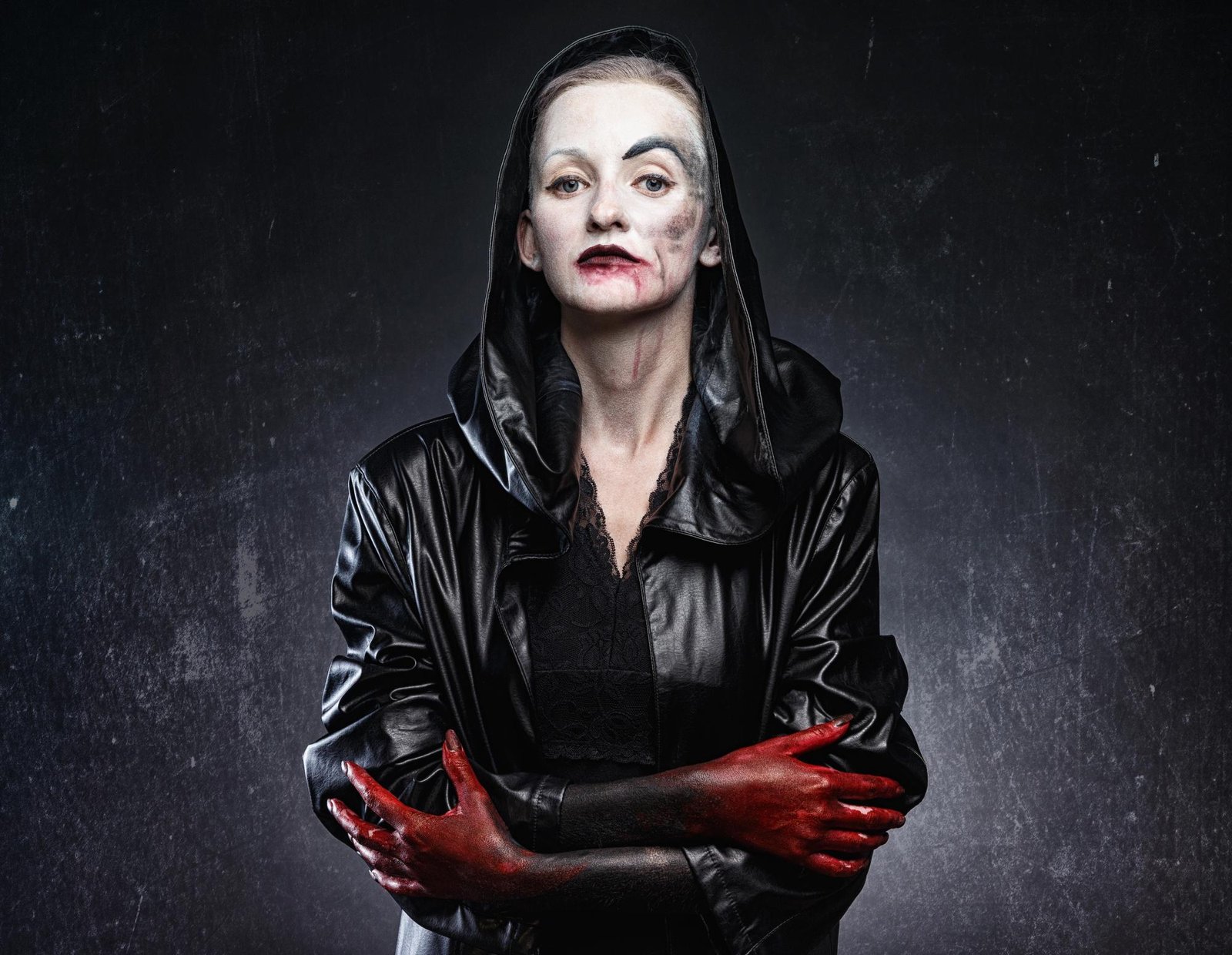 Pale woman in a black hooded coat with blood-smeared hands and gothic makeup, staring directly into camera.