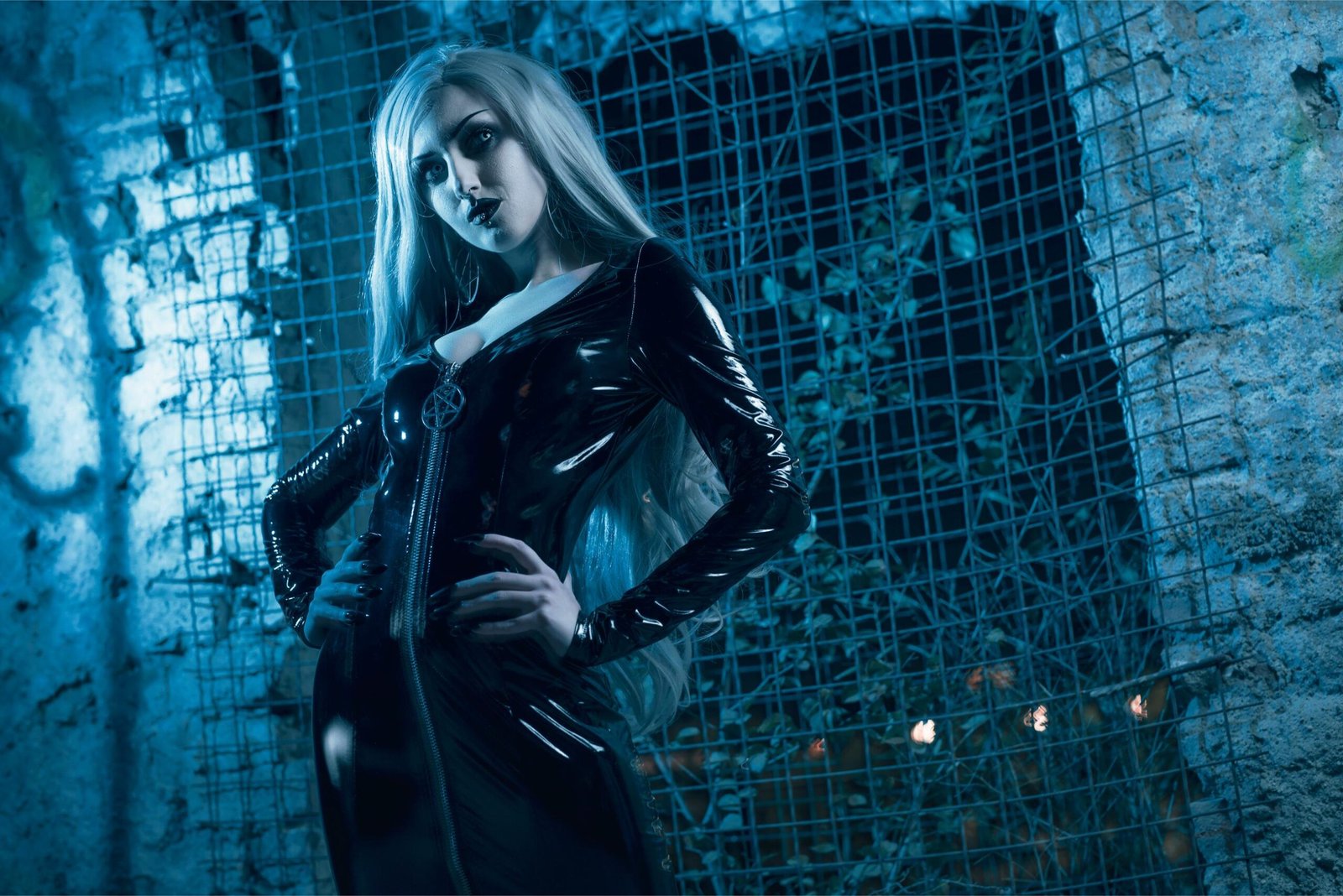 Blonde goth woman in mirror-shine black latex and pentagram pendant, lit by eerie blue in urban gothic setting.