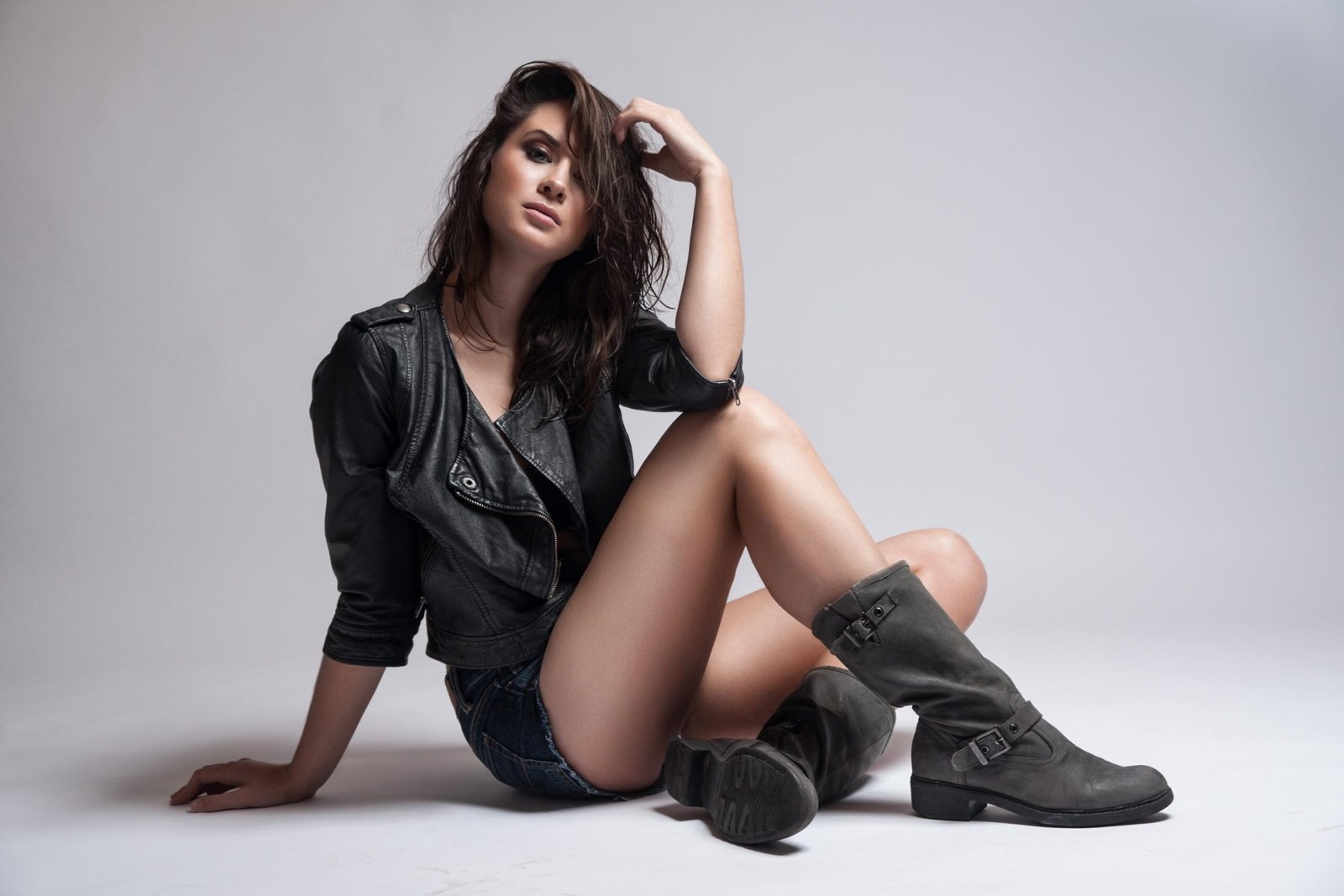 Woman in leather jacket, denim shorts, and boots posing confidently on white studio floor with tousled hair.