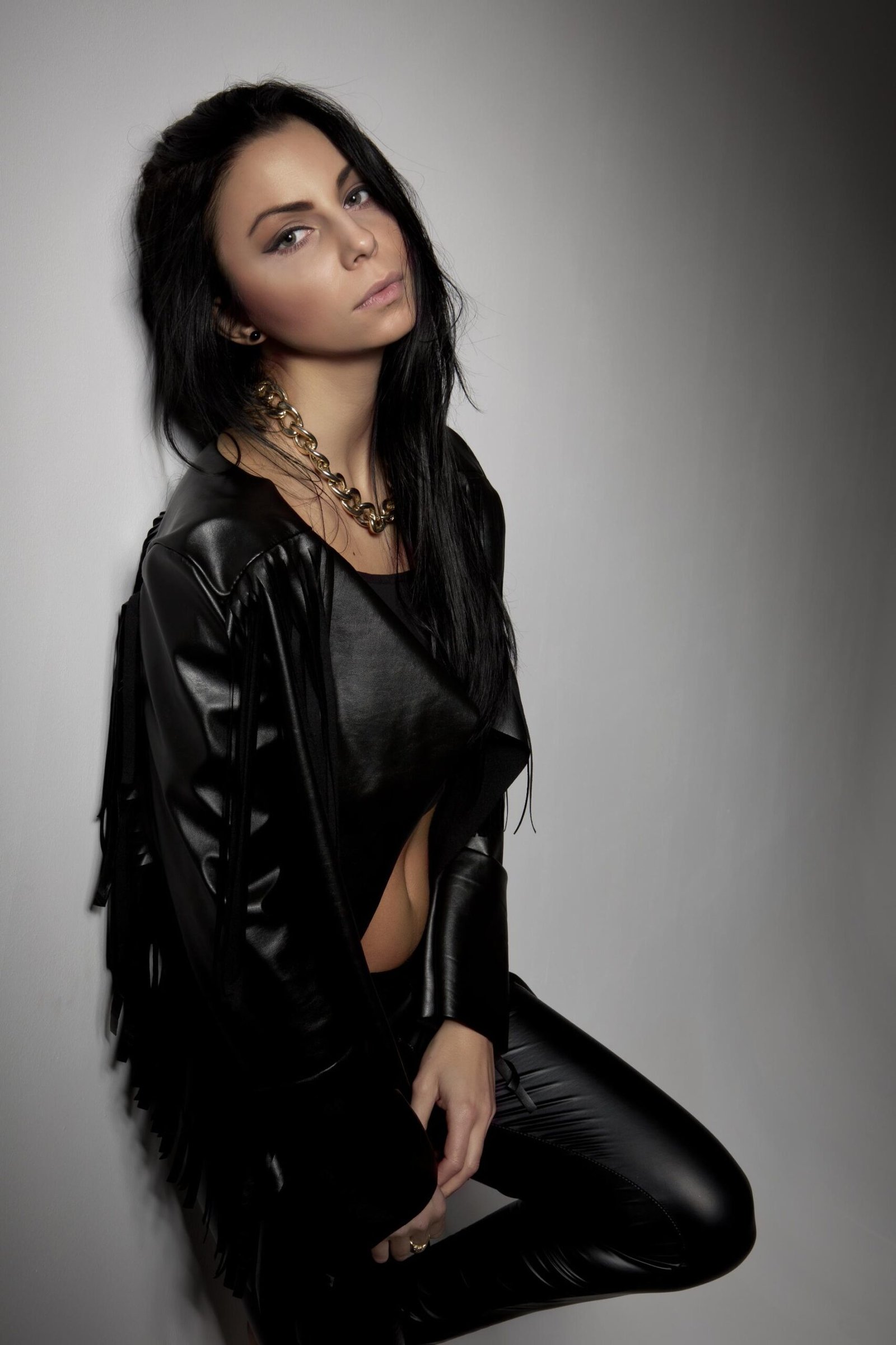 Gothic woman in black leather outfit and gold chain necklace leaning against wall with intense expression.
