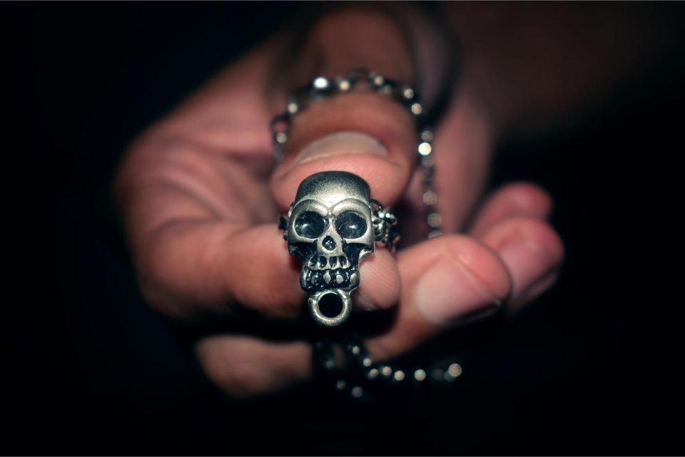 Close-up of a hand holding a silver skull pendant necklace in dramatic low light.