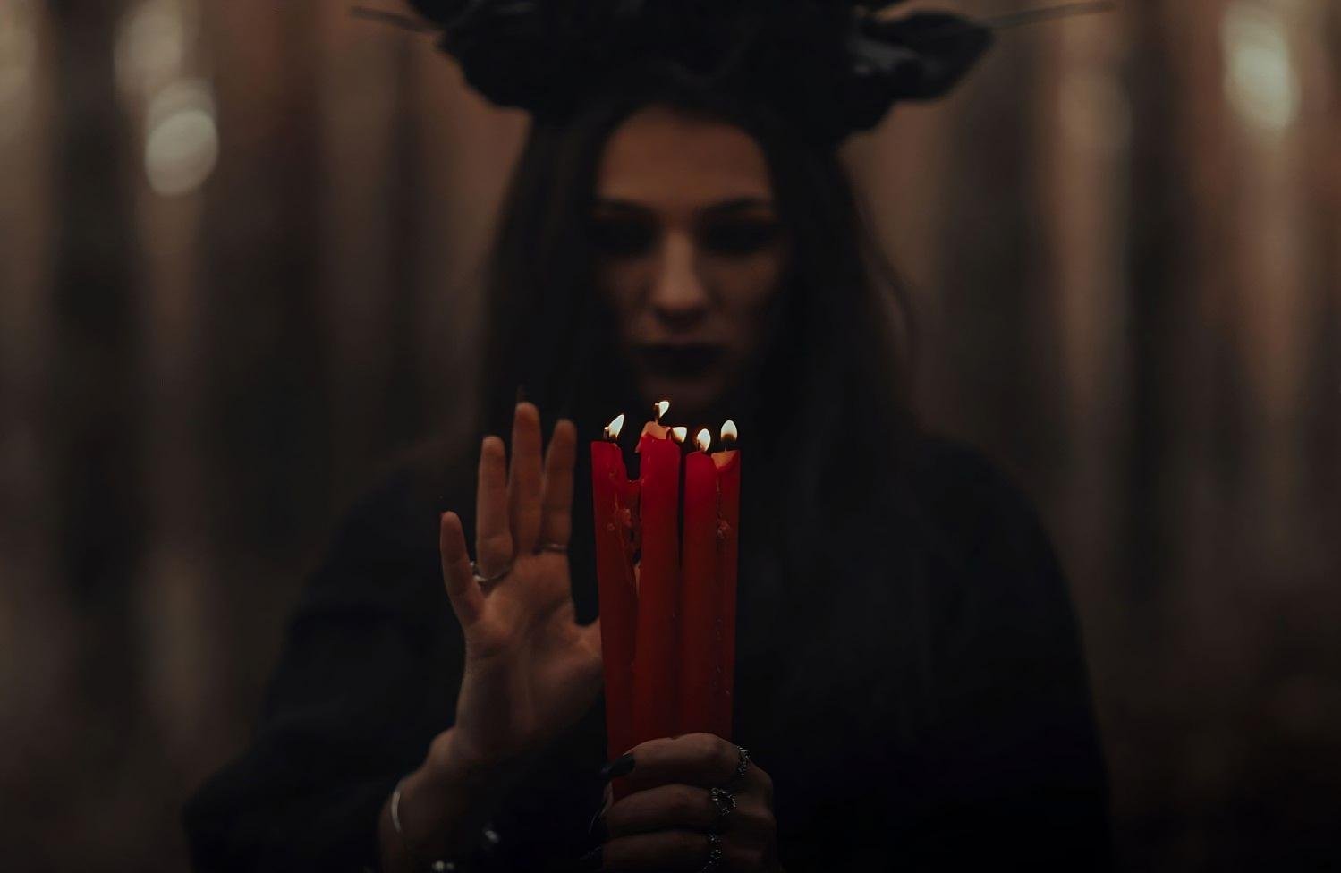 Mysterious goth woman with dark floral crown, holding burning red candles in a misty forest at dusk.