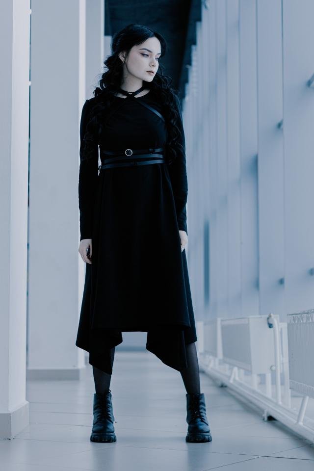 Gothic woman in long black dress and boots standing in a cold-toned hallway, gazing to the side with intensity.