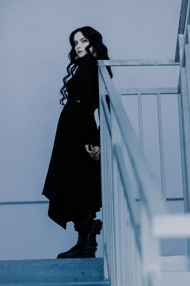 Woman in gothic outfit standing on stairwell, turning back with fierce gaze in a moody, blue-lit scene.