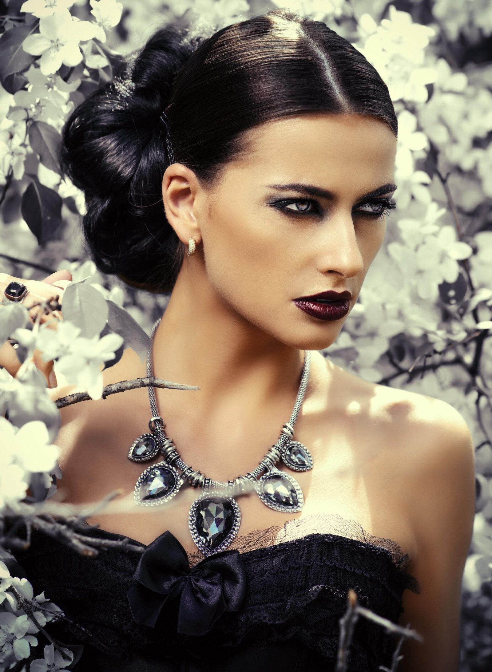 Gothic woman with dark makeup and jeweled necklace wearing a black lace corset among pale flowers.