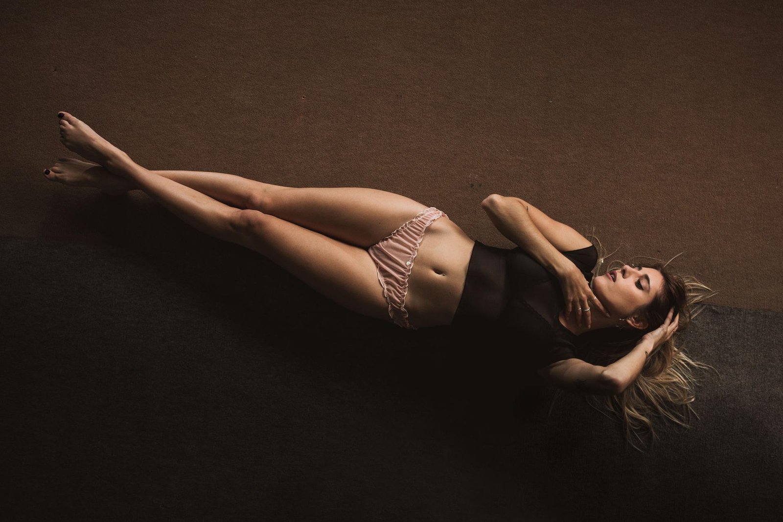 Woman lying flat on a dark floor, wearing a sheer black top and pink ruffled panties, eyes closed. 