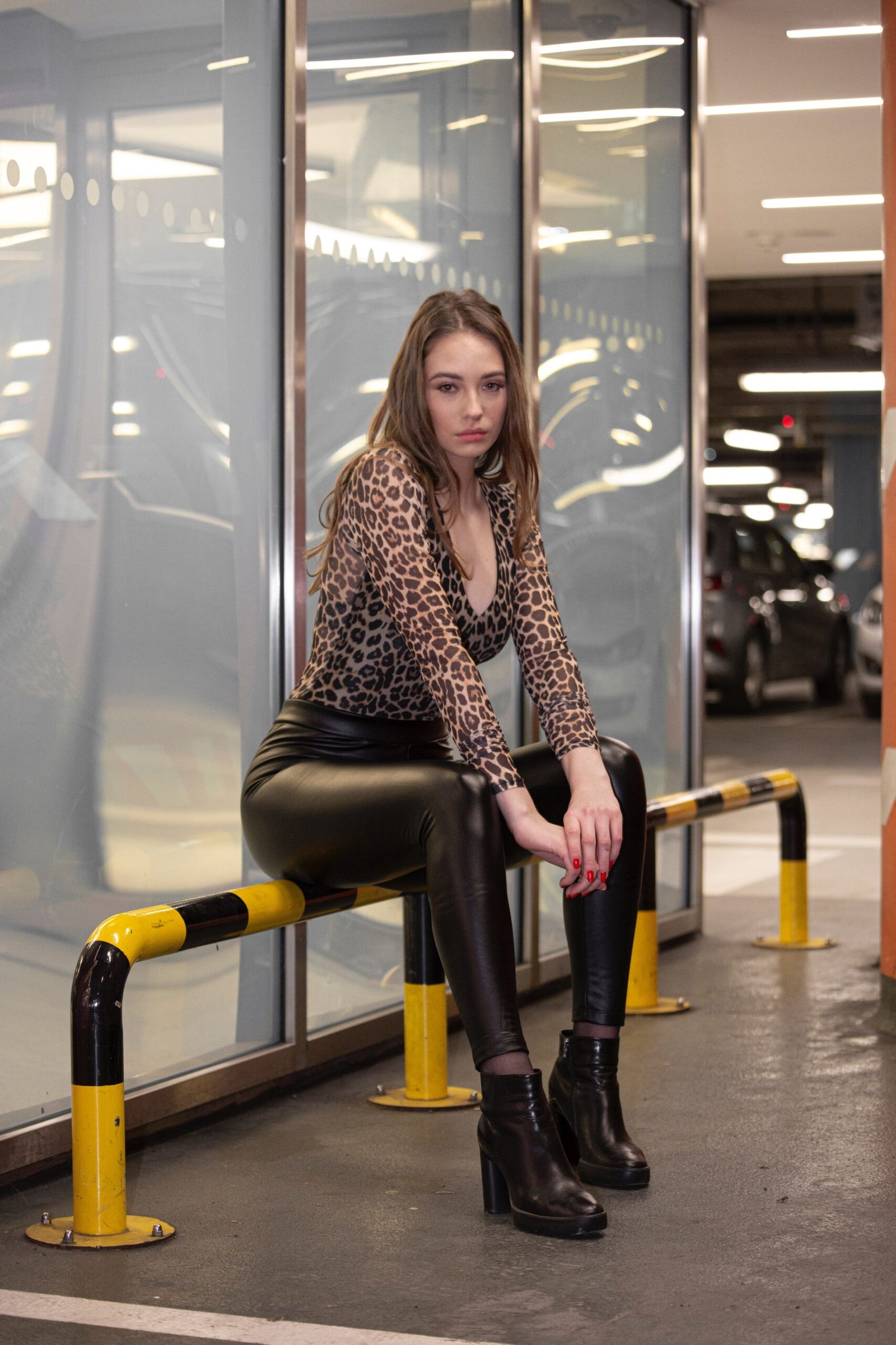 Woman in leopard print top and leather pants sitting on a barrier in a parking garage, wearing black platform boots.