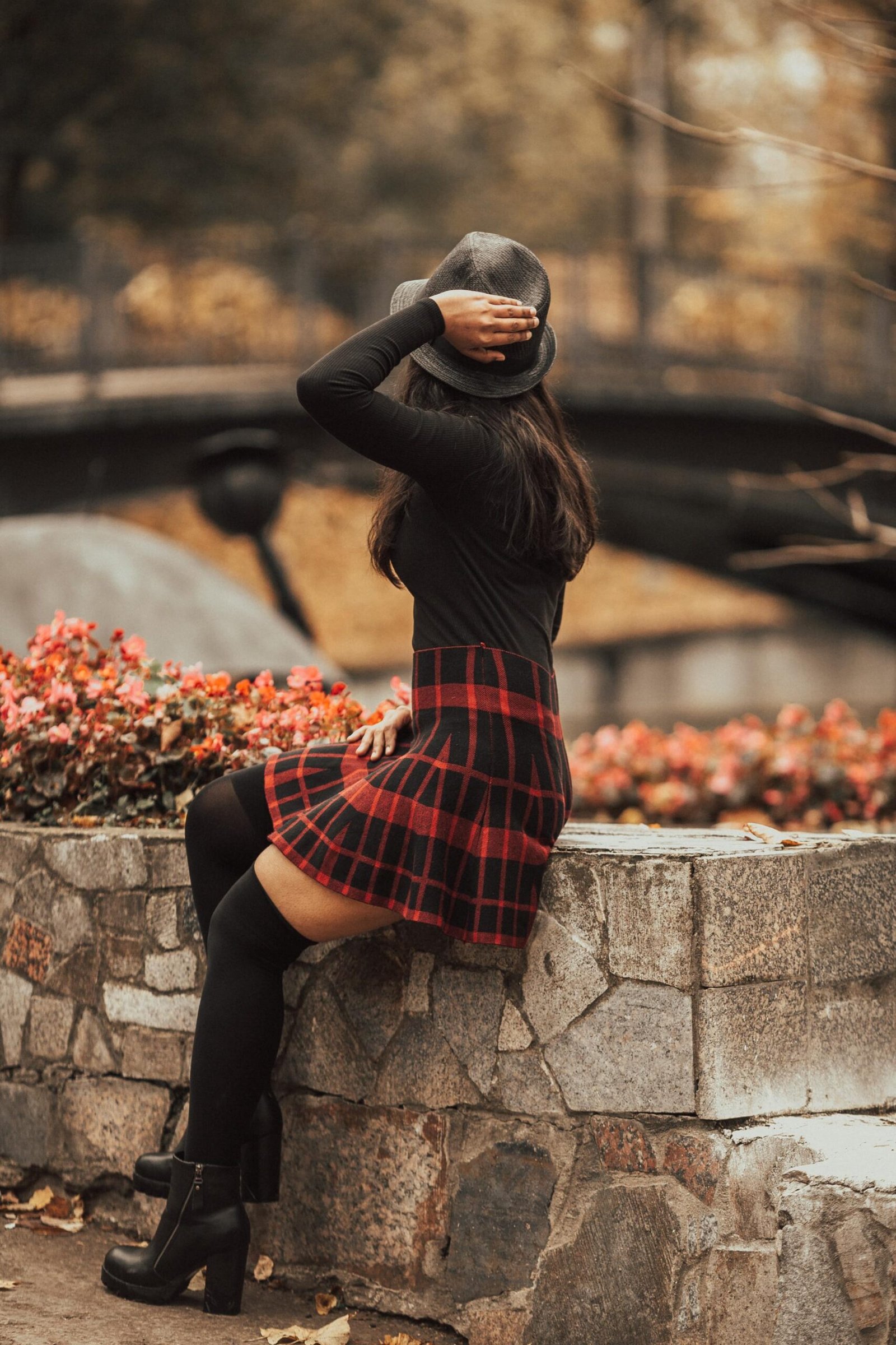 Woman in red plaid skirt and black hat sitting on stone wall in autumn park