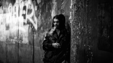 Man with long hair smoking in a dark alley against a graffiti-covered wall