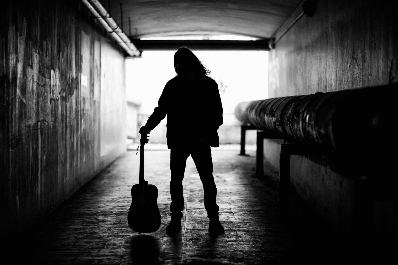 Silhouette of a person holding a guitar in a dark tunnel, captured in harsh black-and-white lighting that emphasizes metallic solitude.