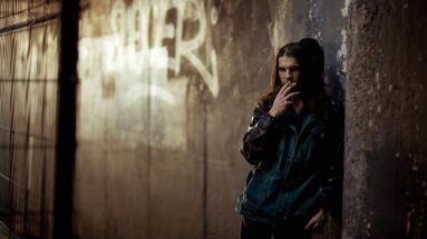 Man with long hair smokes a cigarette against a graffiti wall in a dim urban tunnel.