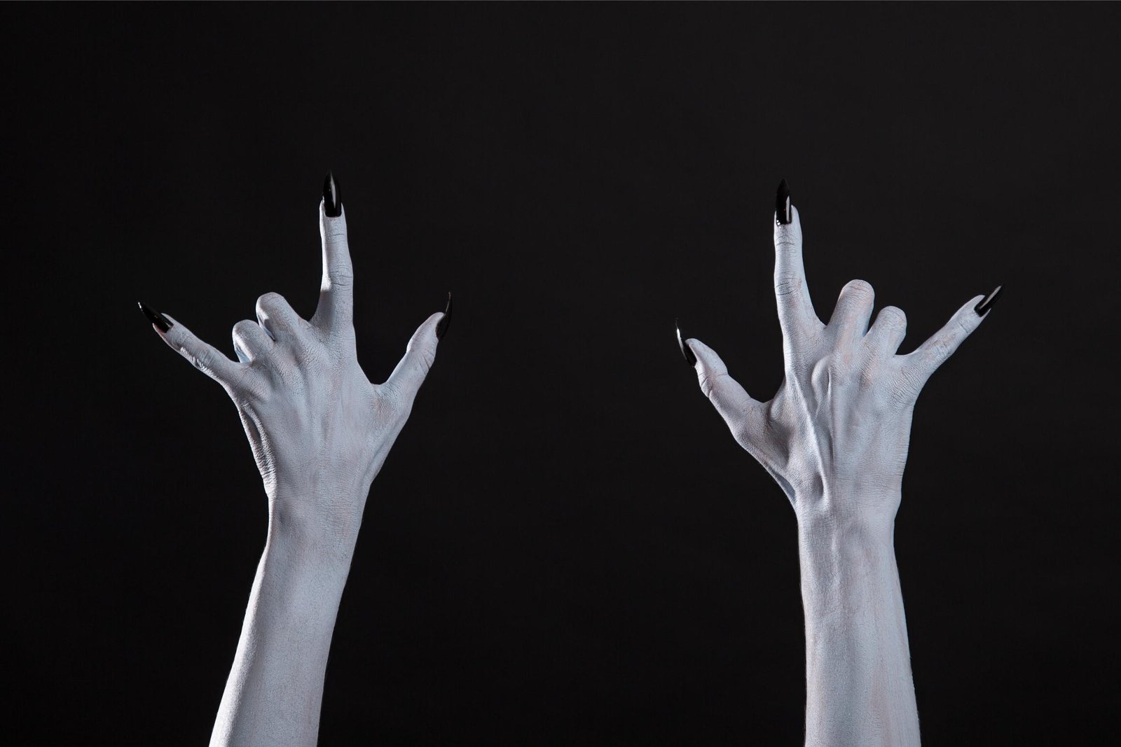 Two pale hands with long black nails raised in devil horns against black background