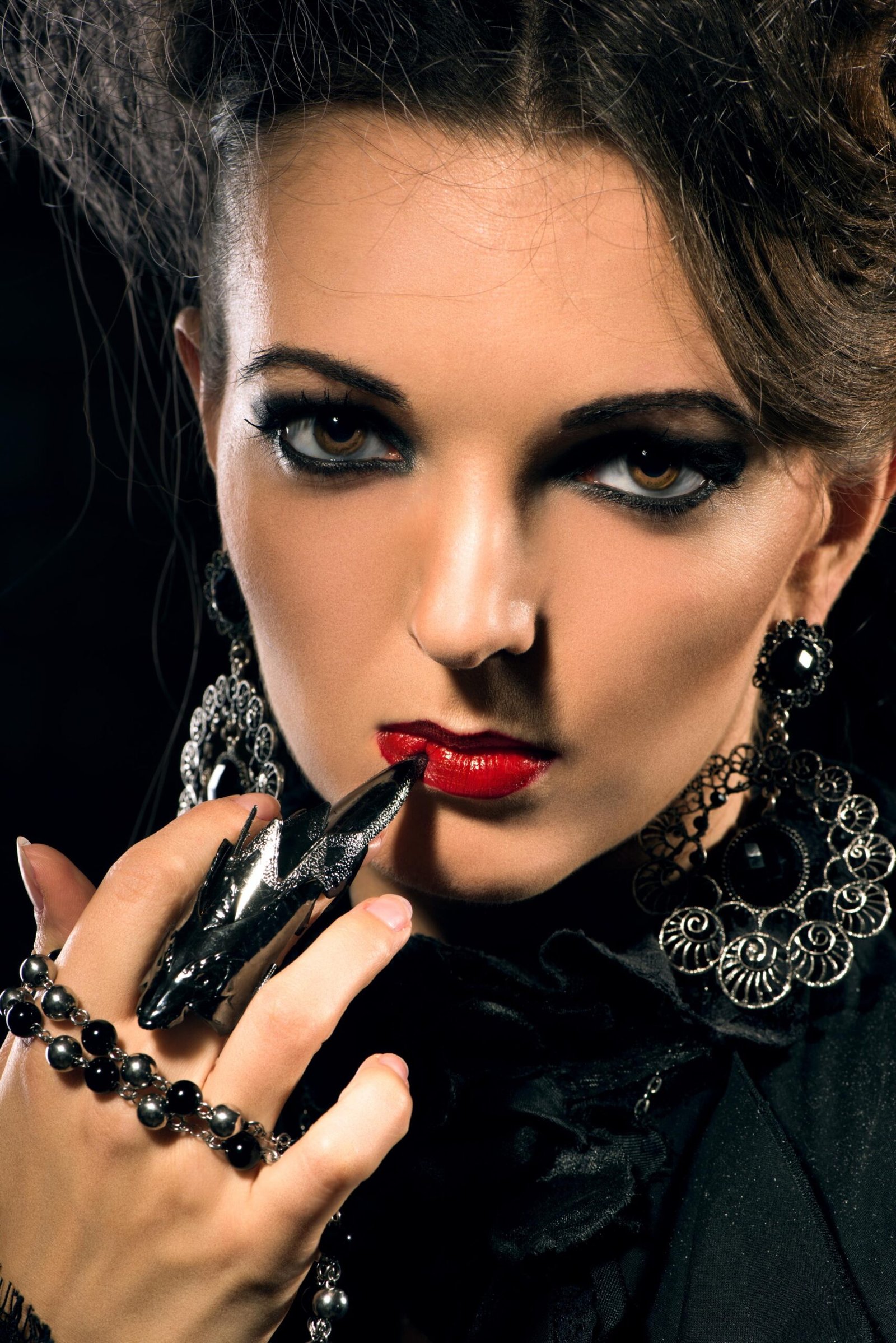 Close-up of woman with red lips, gothic jewelry and dramatic black eyeliner holding finger to mouth