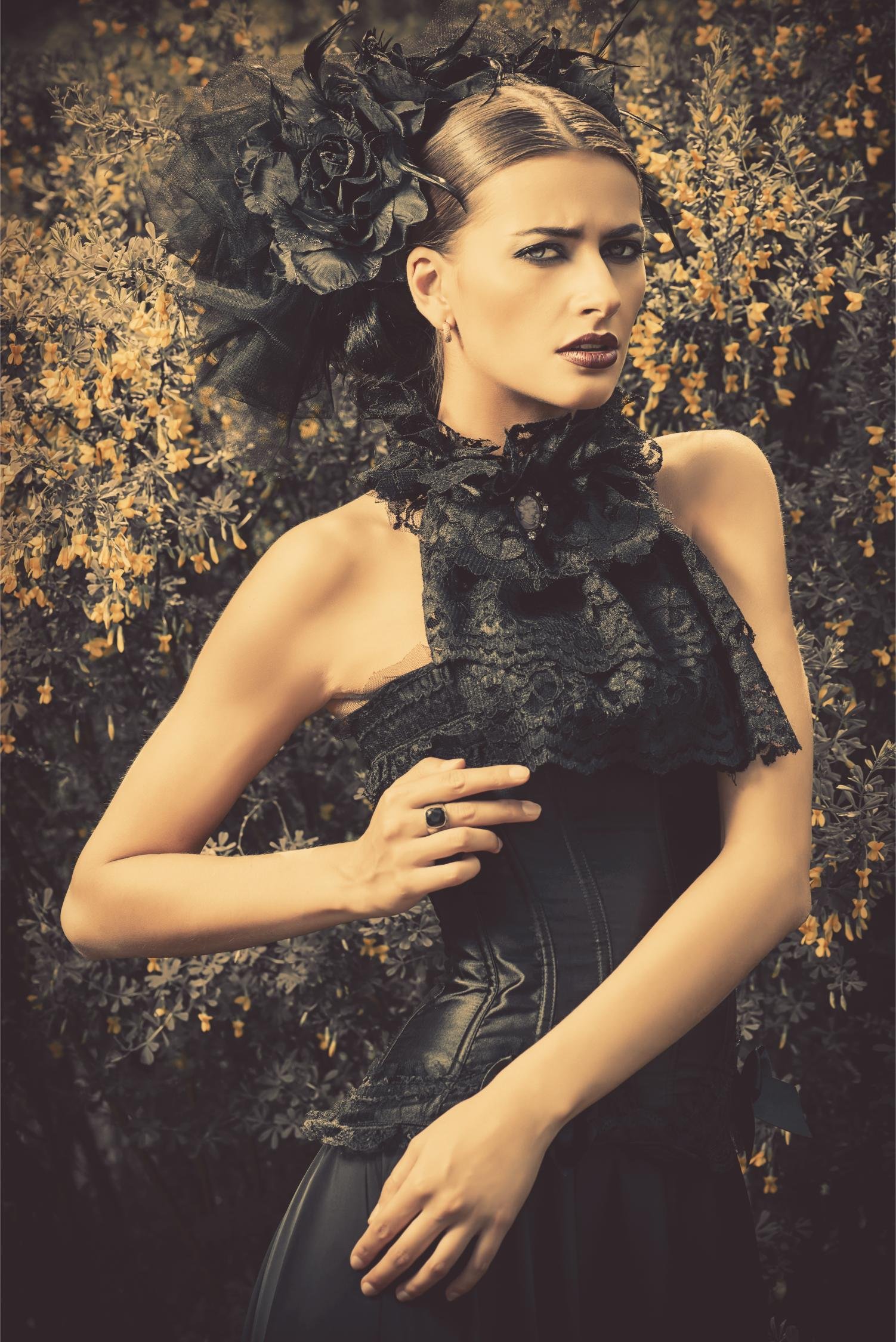 Gothic woman in black lace corset and large floral headpiece posing against yellow flowering bushes.