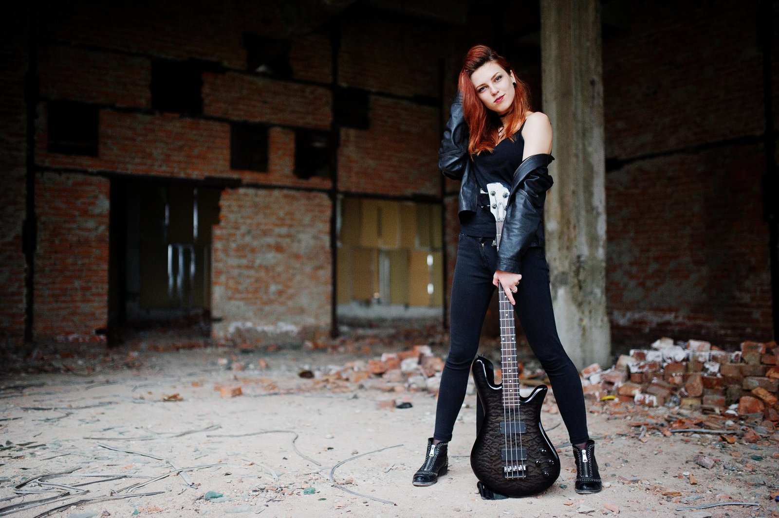 Woman in black leather jacket posing with bass guitar in abandoned building