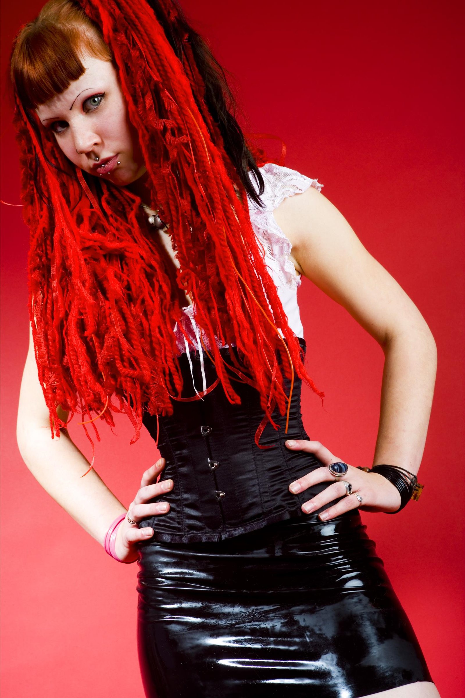 Gothic woman with red dreadlocks in corset and black latex skirt against red backdrop