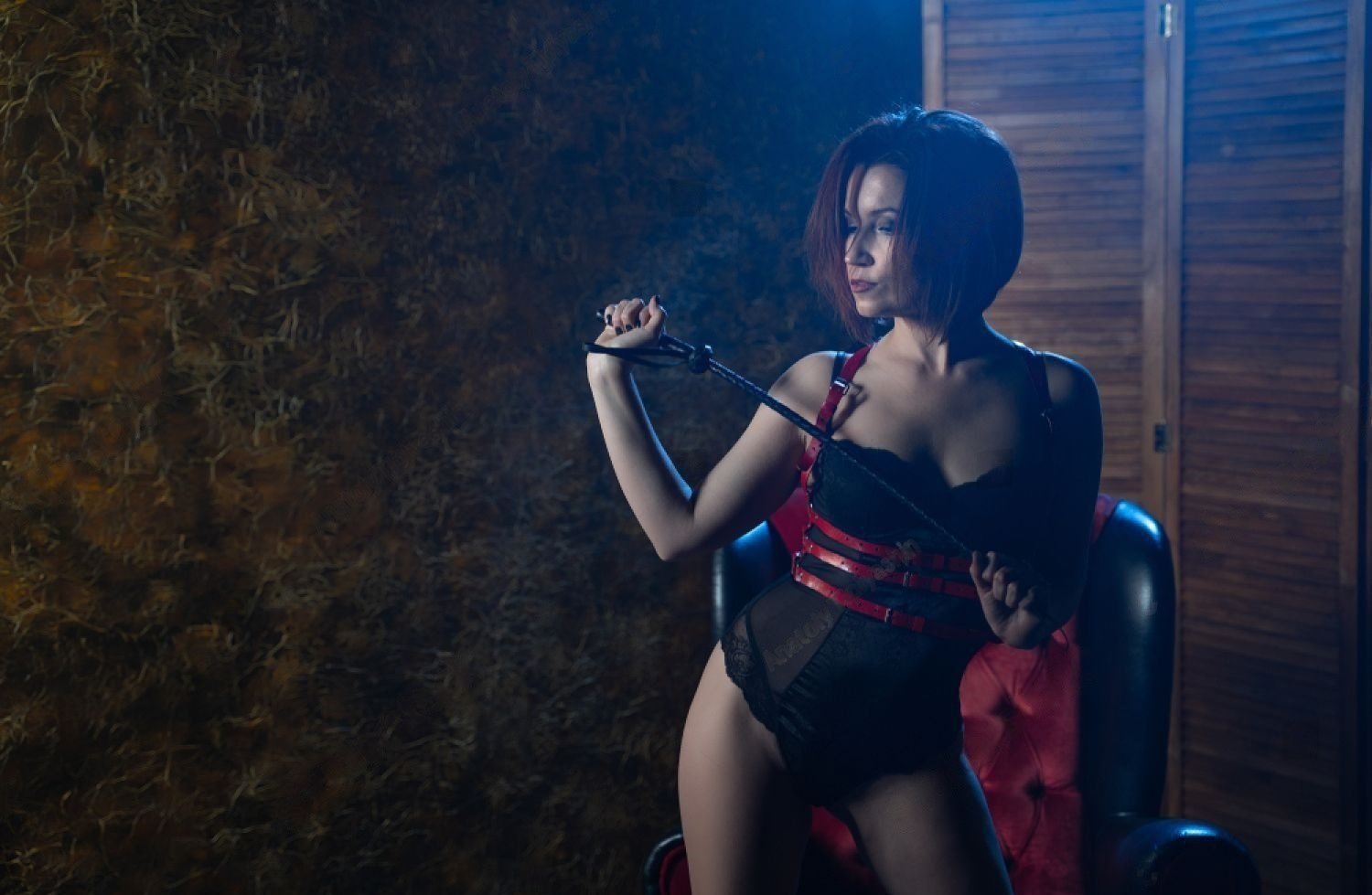  Woman with short red hair in black and red bondage gear holding a whip, posing with dominant attitude in cool blue stage light.