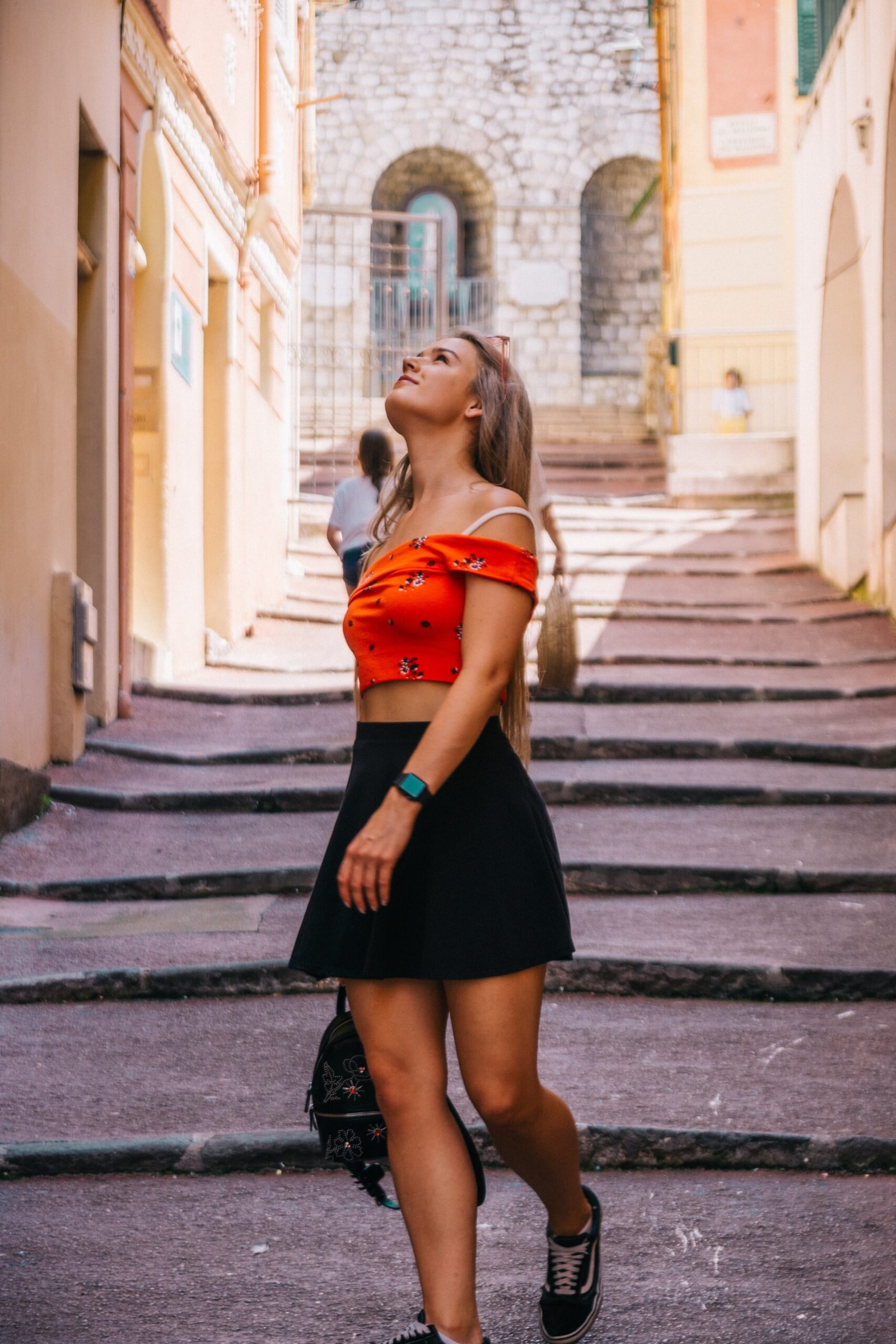 Young woman in a red crop top and black skirt walking up a sunlit stone path in an old town, gazing upward with confidence.