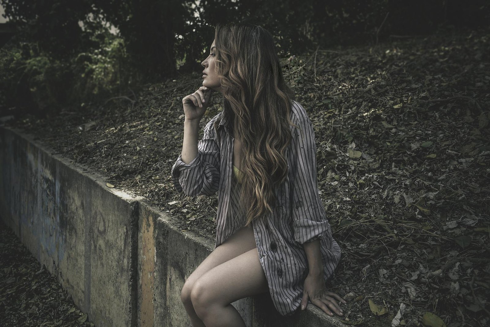 Woman in an oversized shirt sitting by a concrete ledge surrounded by leaves, with a dreamy yet dark natural expression.