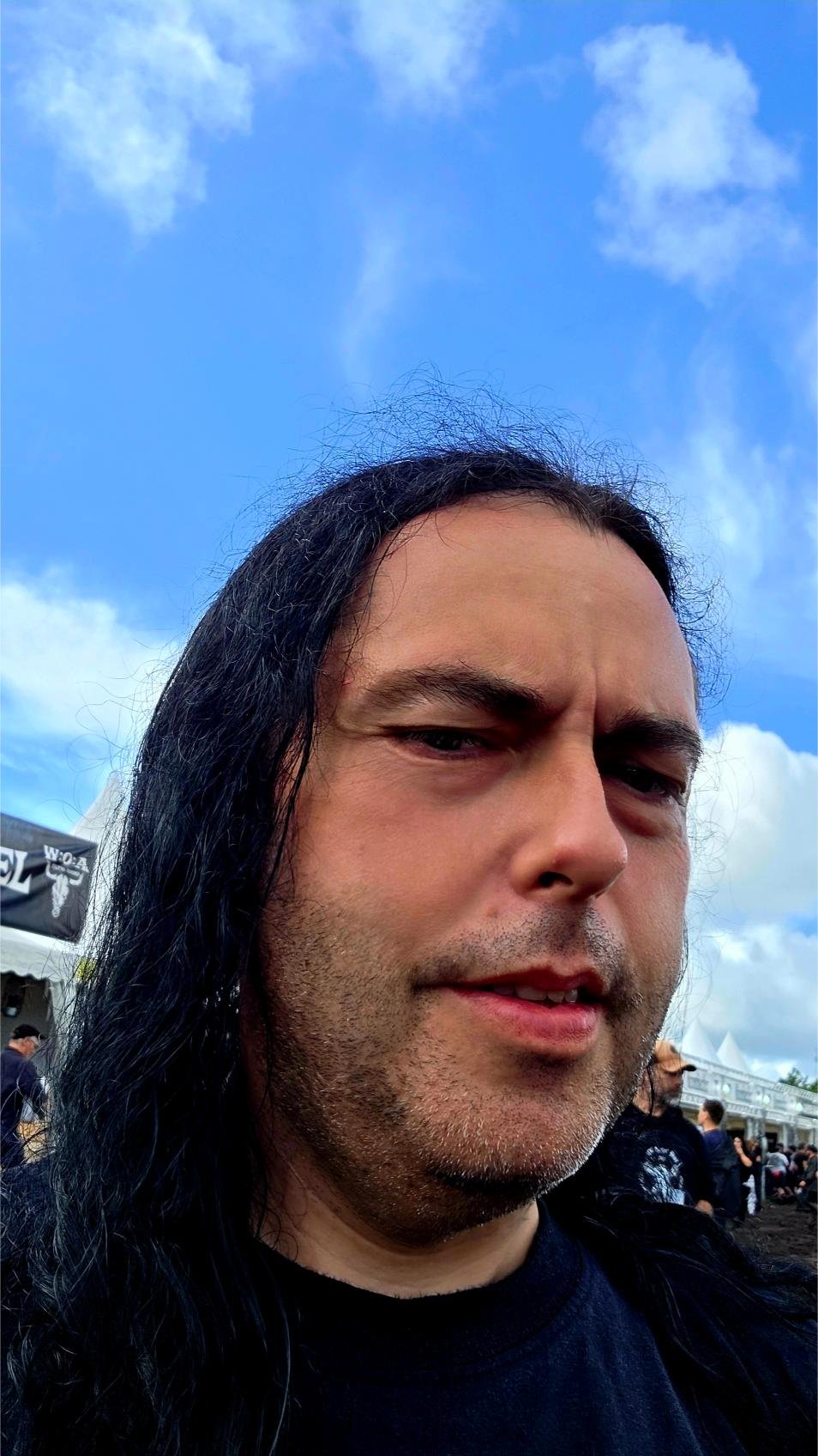 Xavi at Wacken Open Air 2025, long black hair and black metal shirt under a blue sky