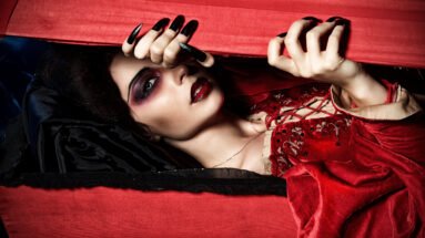 Gothic woman in red velvet dress lying inside coffin with dramatic makeup.