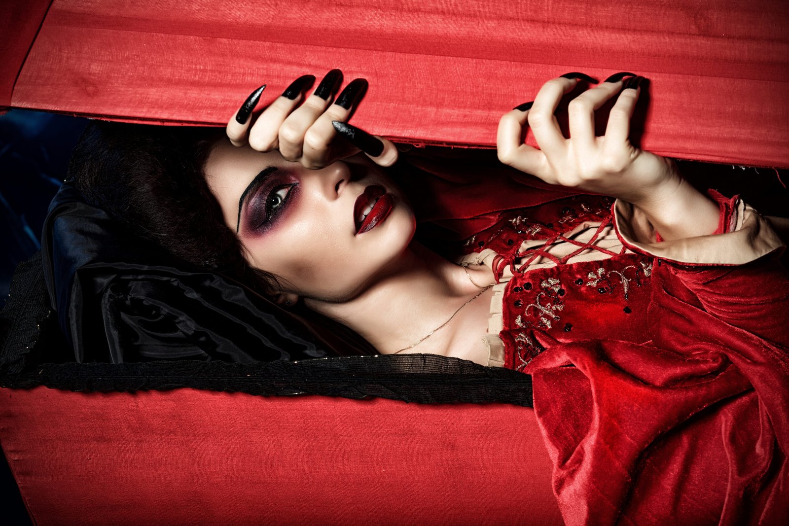 Gothic woman in red velvet dress lying inside coffin with dramatic makeup.