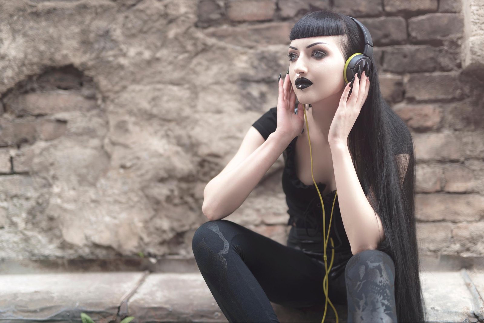 Goth woman with headphones sitting by a stone wall, wearing black makeup and long black hair