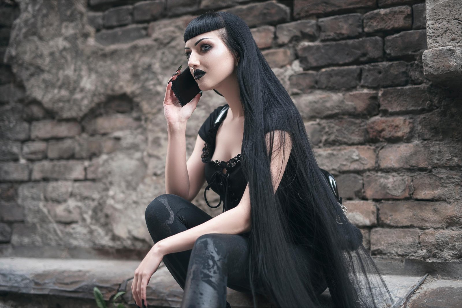 Goth woman in black clothes talking on a smartphone against a brick wall