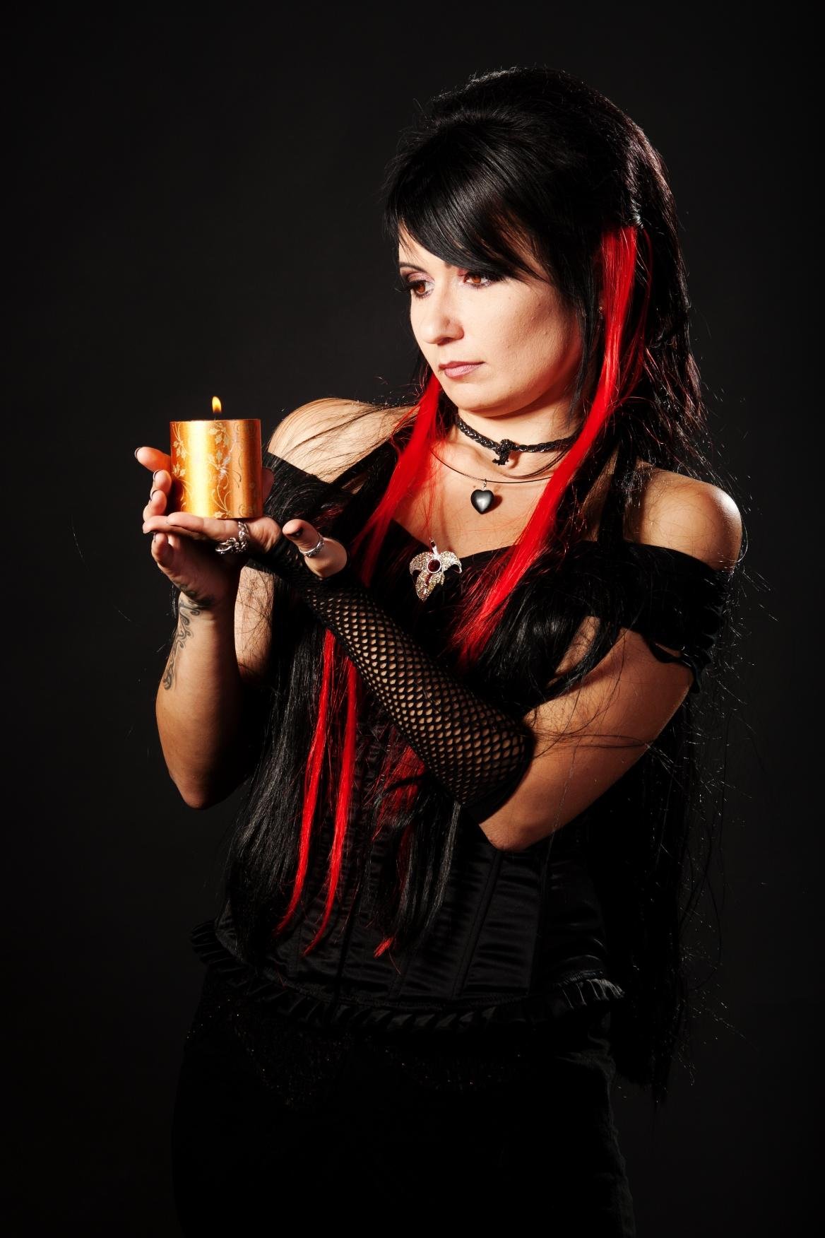Gothic woman with long black and red hair holding a lit candle, wearing a black corset and fishnet gloves.