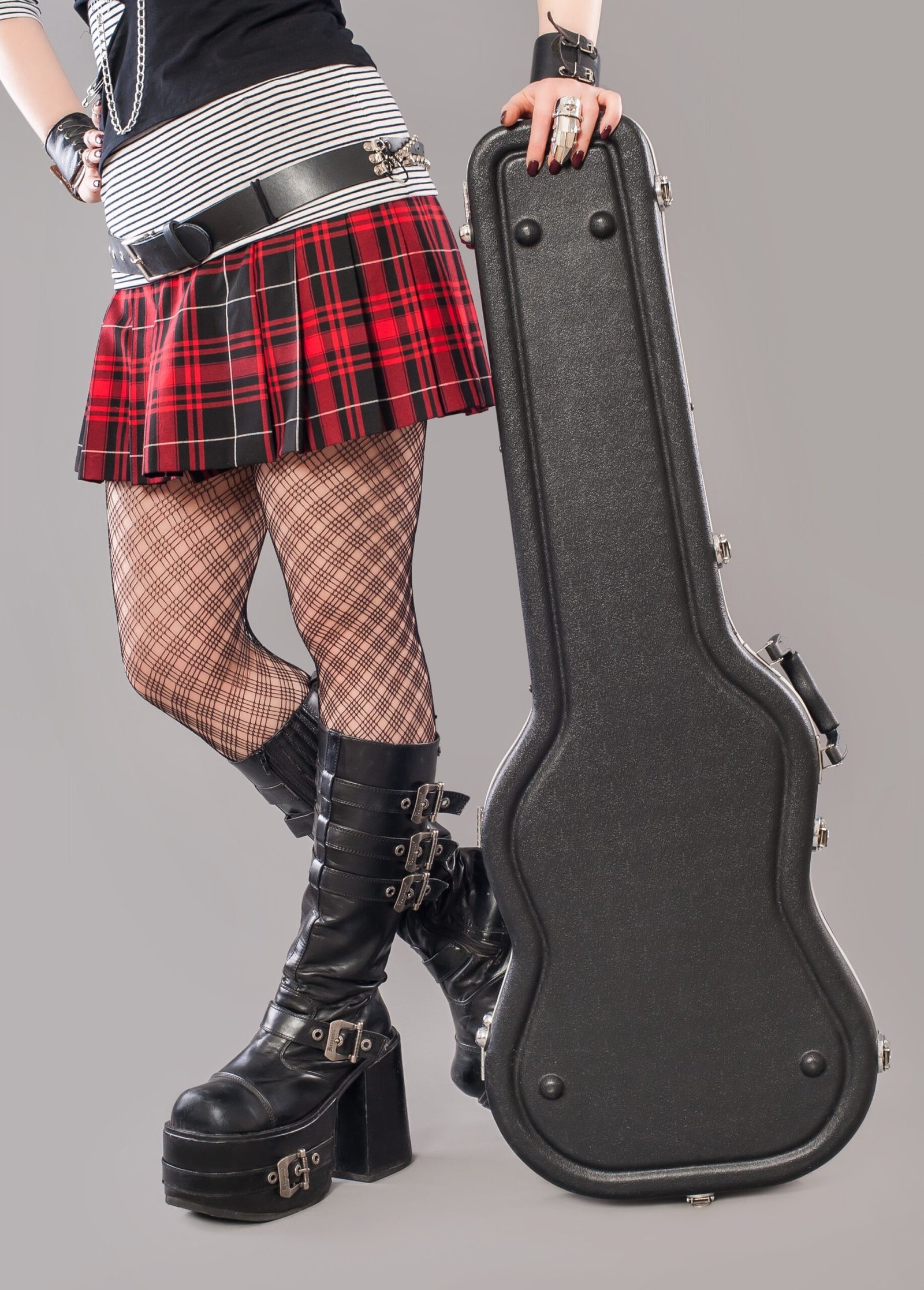 A person in a black and white striped shirt and a red plaid mini skirt, with fishnet tights and tall platform boots, holding a guitar case.