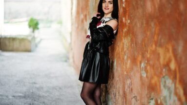 A person leaning against a textured brown wall, wearing a black leather jacket, a printed top, and a black pleated mini skirt.