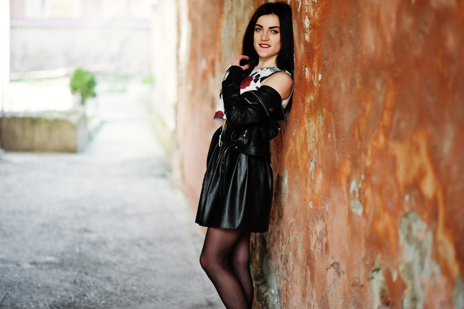 A person leaning against a textured brown wall, wearing a black leather jacket, a printed top, and a black pleated mini skirt.