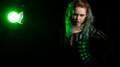 Gothic woman with green-highlighted hair wearing a black corset outfit under dramatic green stage lighting.