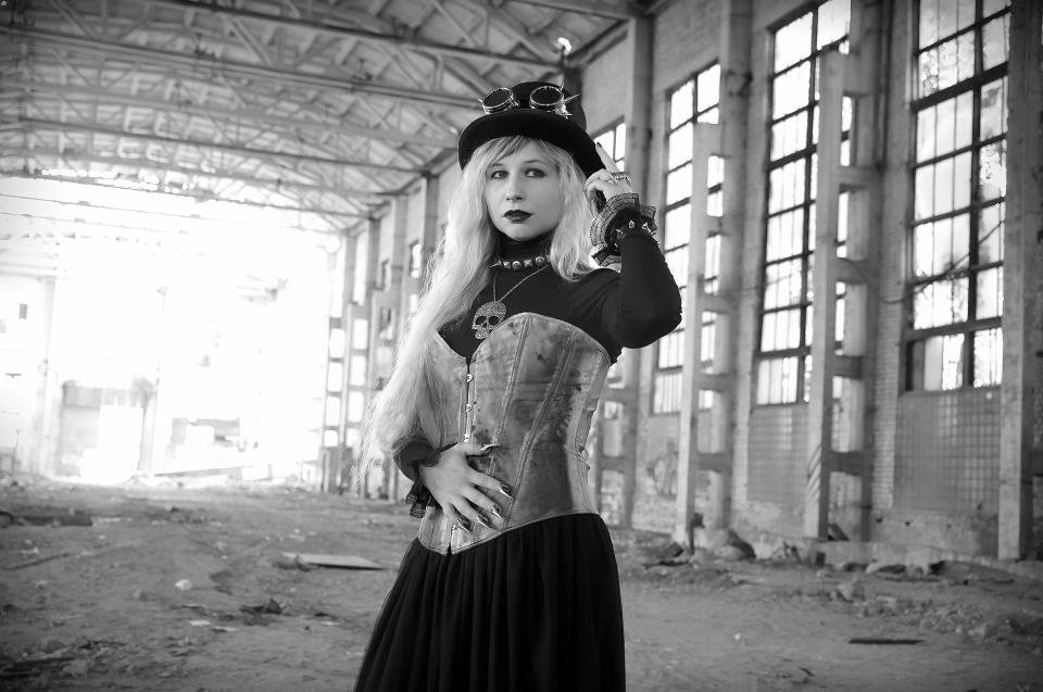 Steampunk woman with long blonde hair in a metallic corset and top hat posing inside an abandoned industrial building.