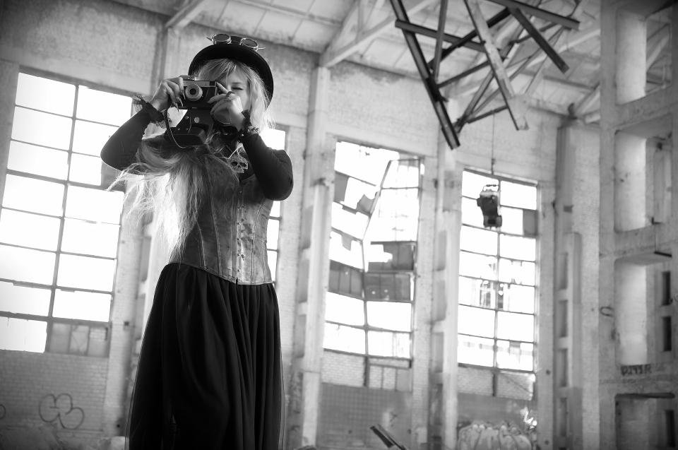 Steampunk woman in corset and top hat photographing with a vintage camera inside a ruined industrial hall.