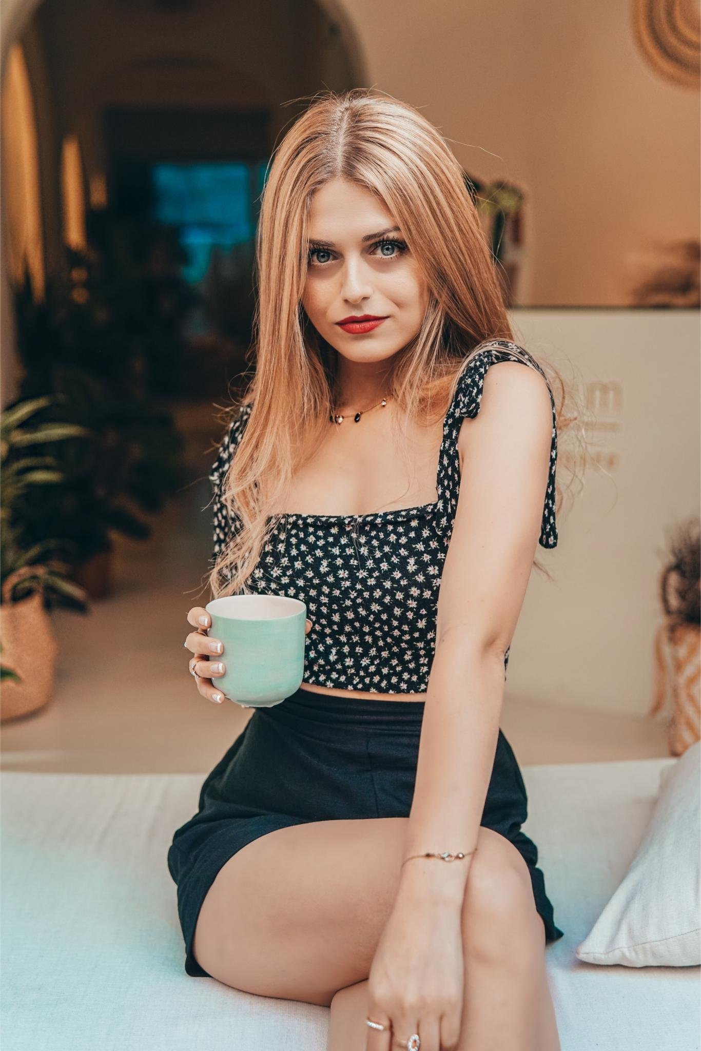 Blonde woman in floral top holding cup, sitting indoors with confident gaze.