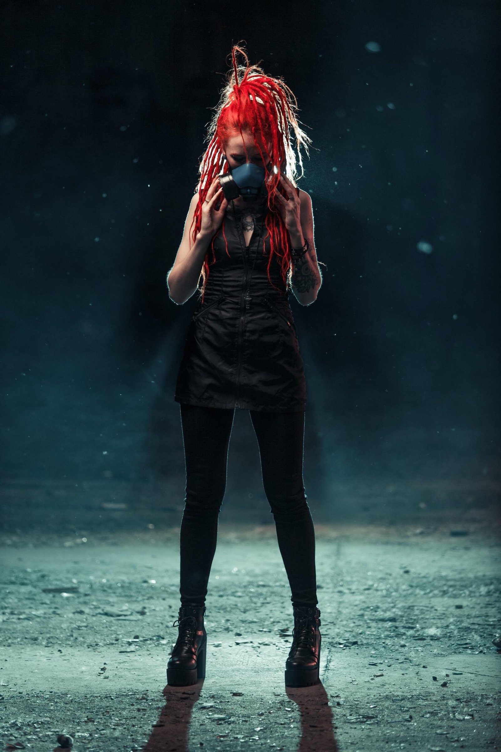 Woman with red dreadlocks wearing gas mask, standing in moody spotlight.