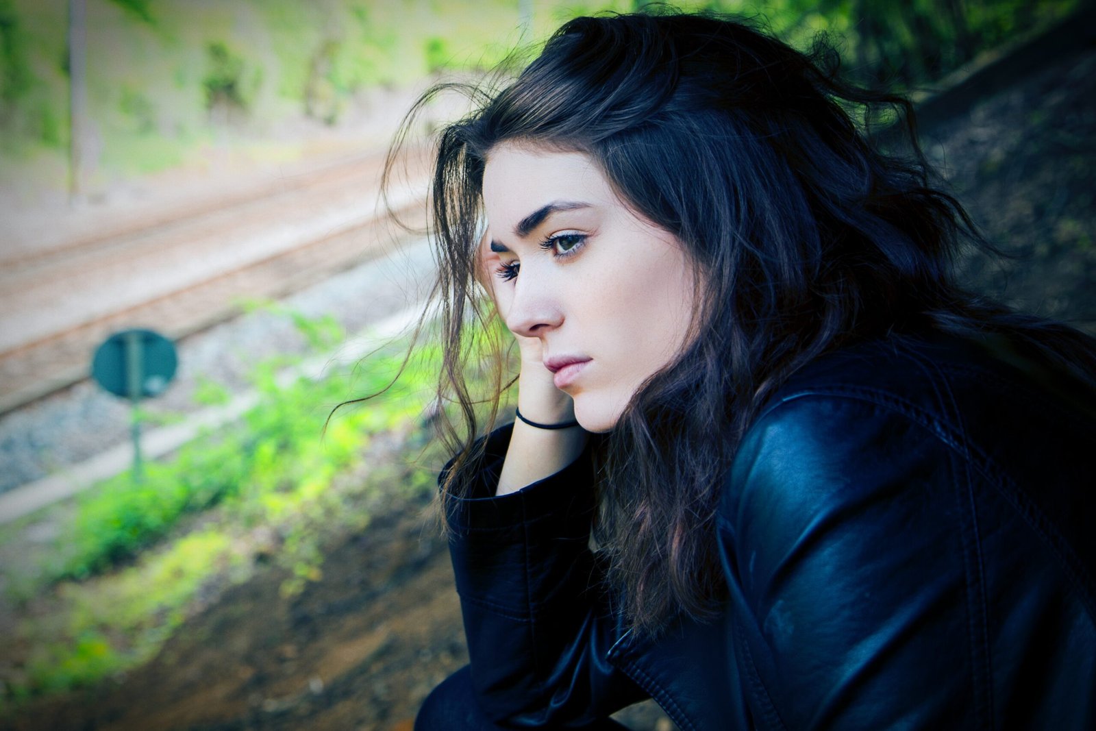 Moody goth woman with dark hair by train tracks, black leather jacket, alternative portrait with deep contrast.