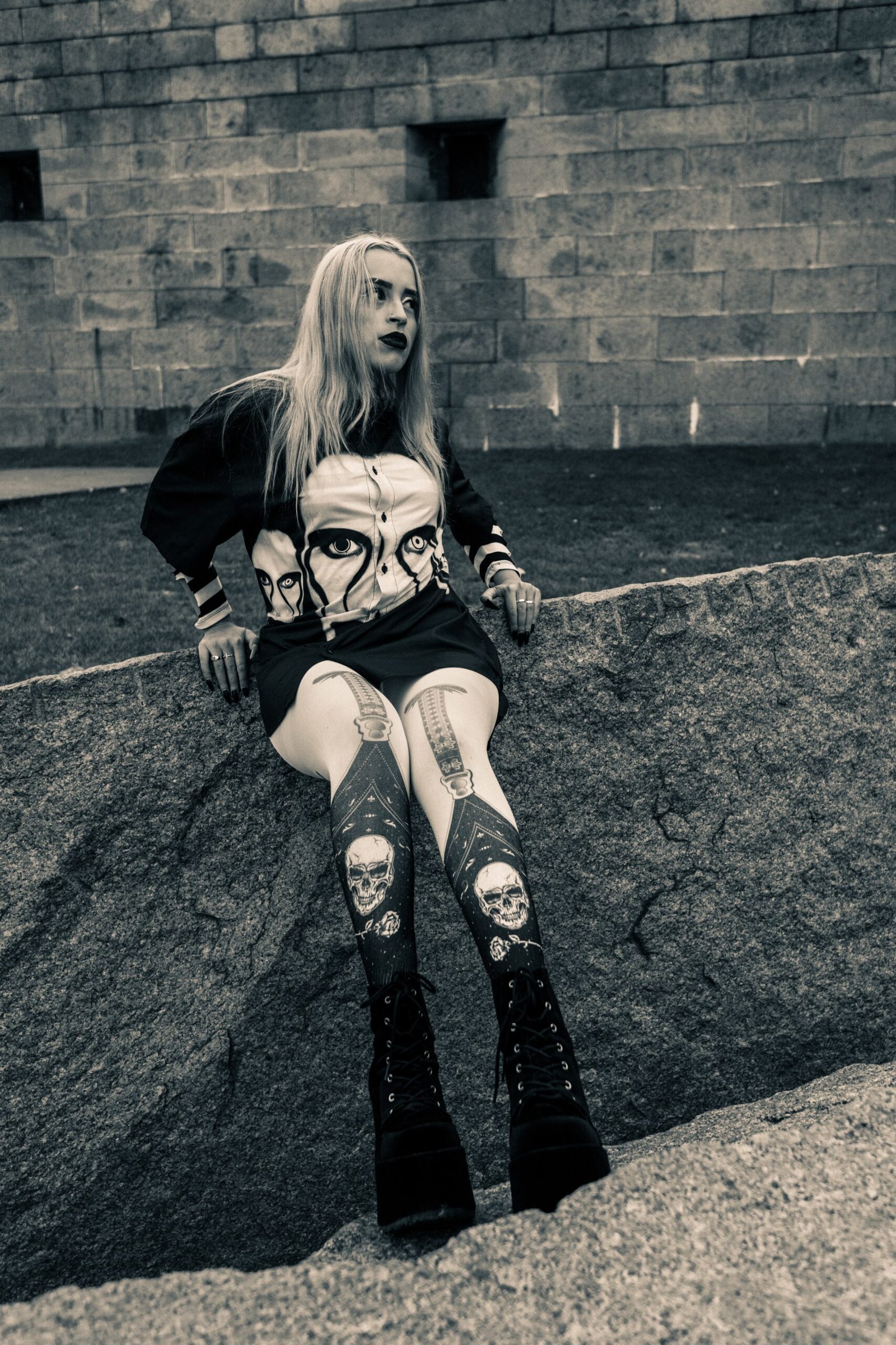 Goth woman in skull-print tights and platform boots, leaning on stone wall, alternative street