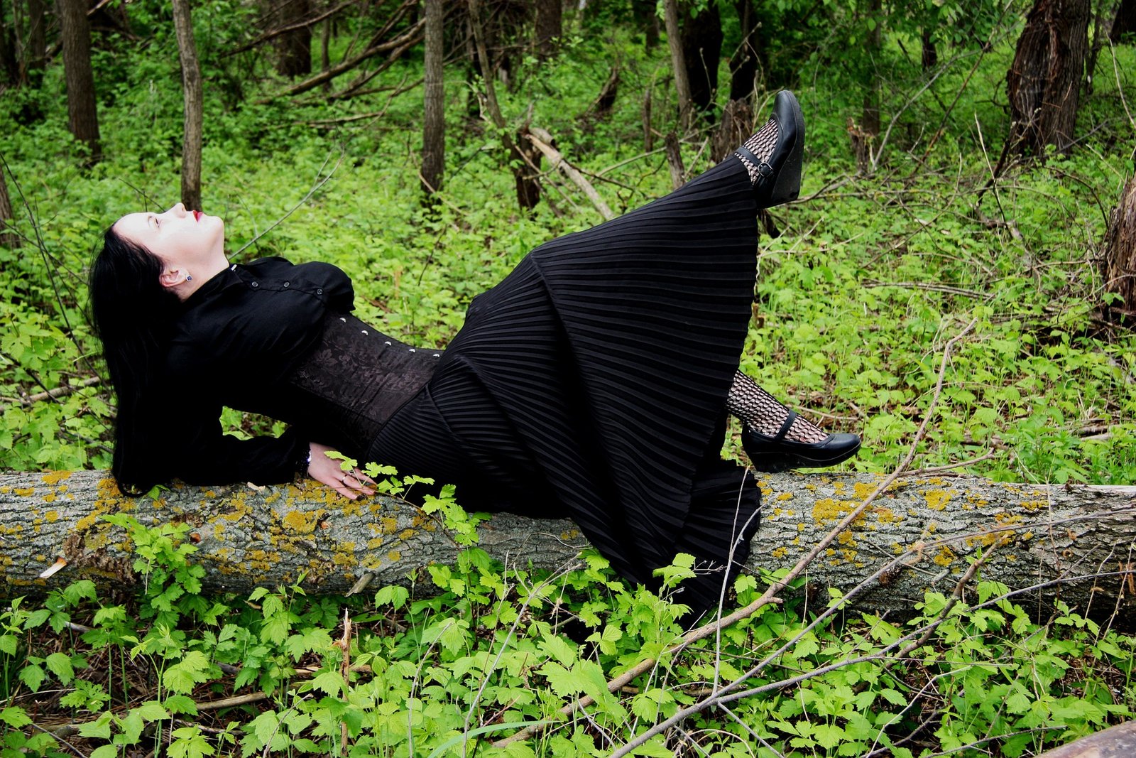 Goth woman kneeling in the woods in a black dress, a corset, and white flowers on her arms.