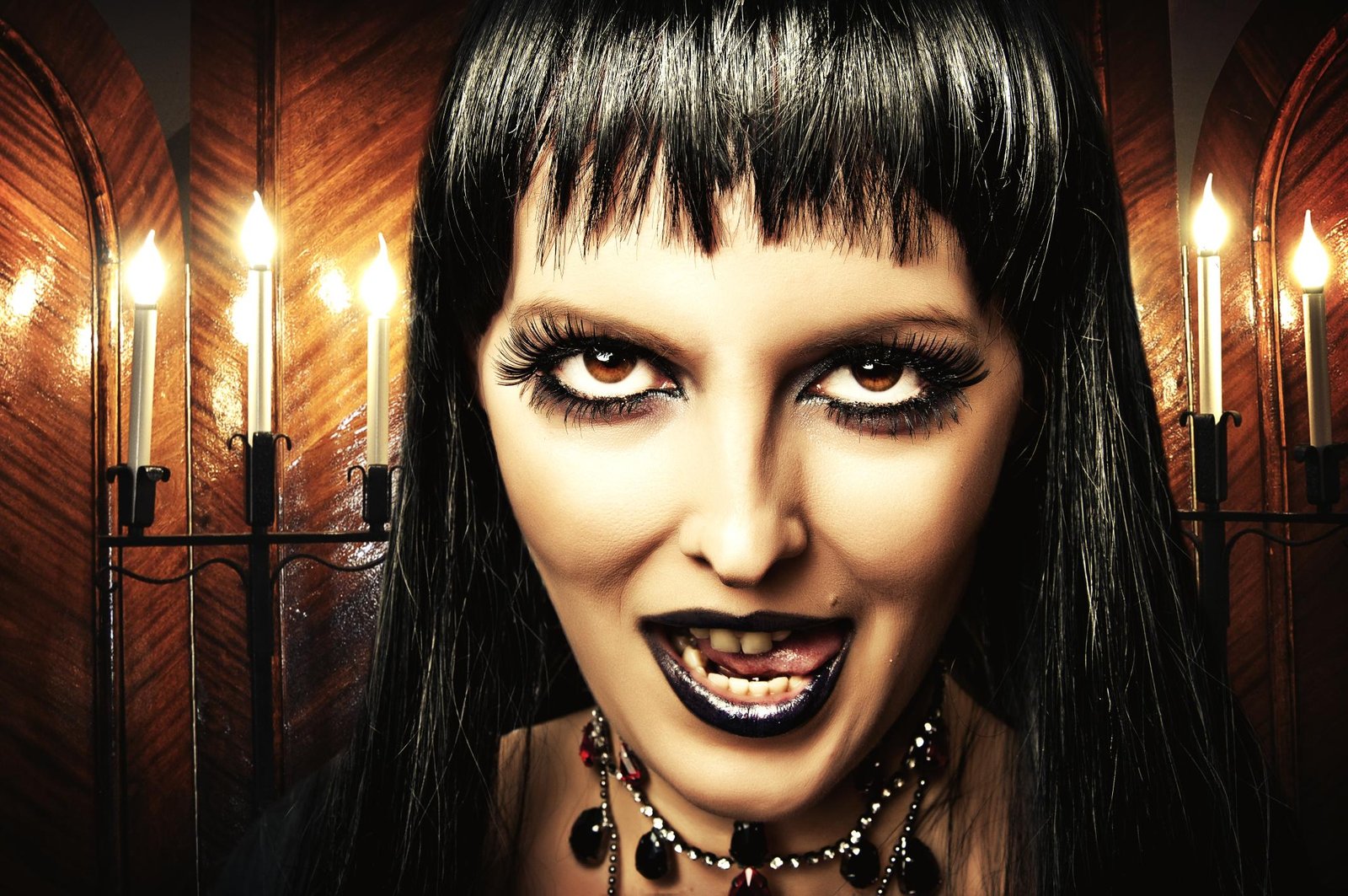 Gothic woman with black hair, dark makeup, and black lipstick smirking in candlelit room.