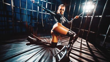 Woman in black latex bodysuit and thigh-high boots holding chains in dark room.