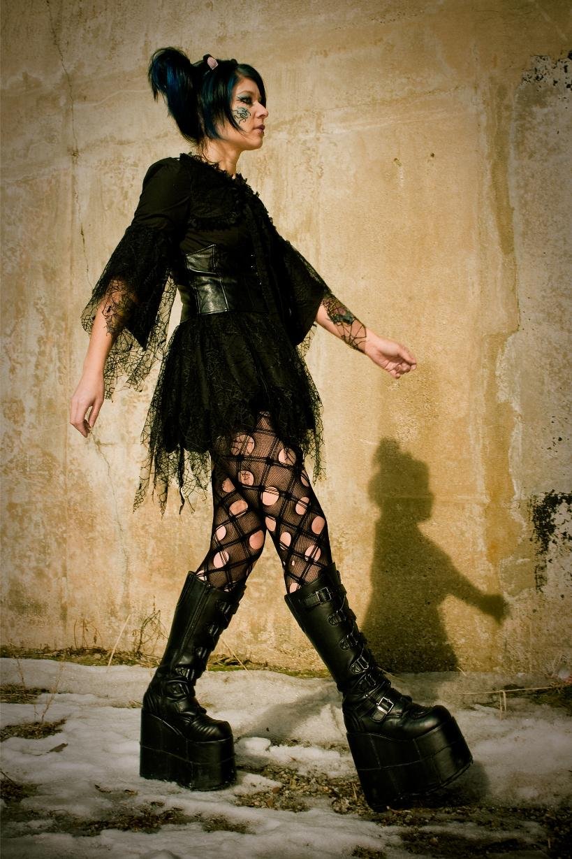 Cyber goth woman in platform boots, black lace skirt, and torn fishnets walking by wall.