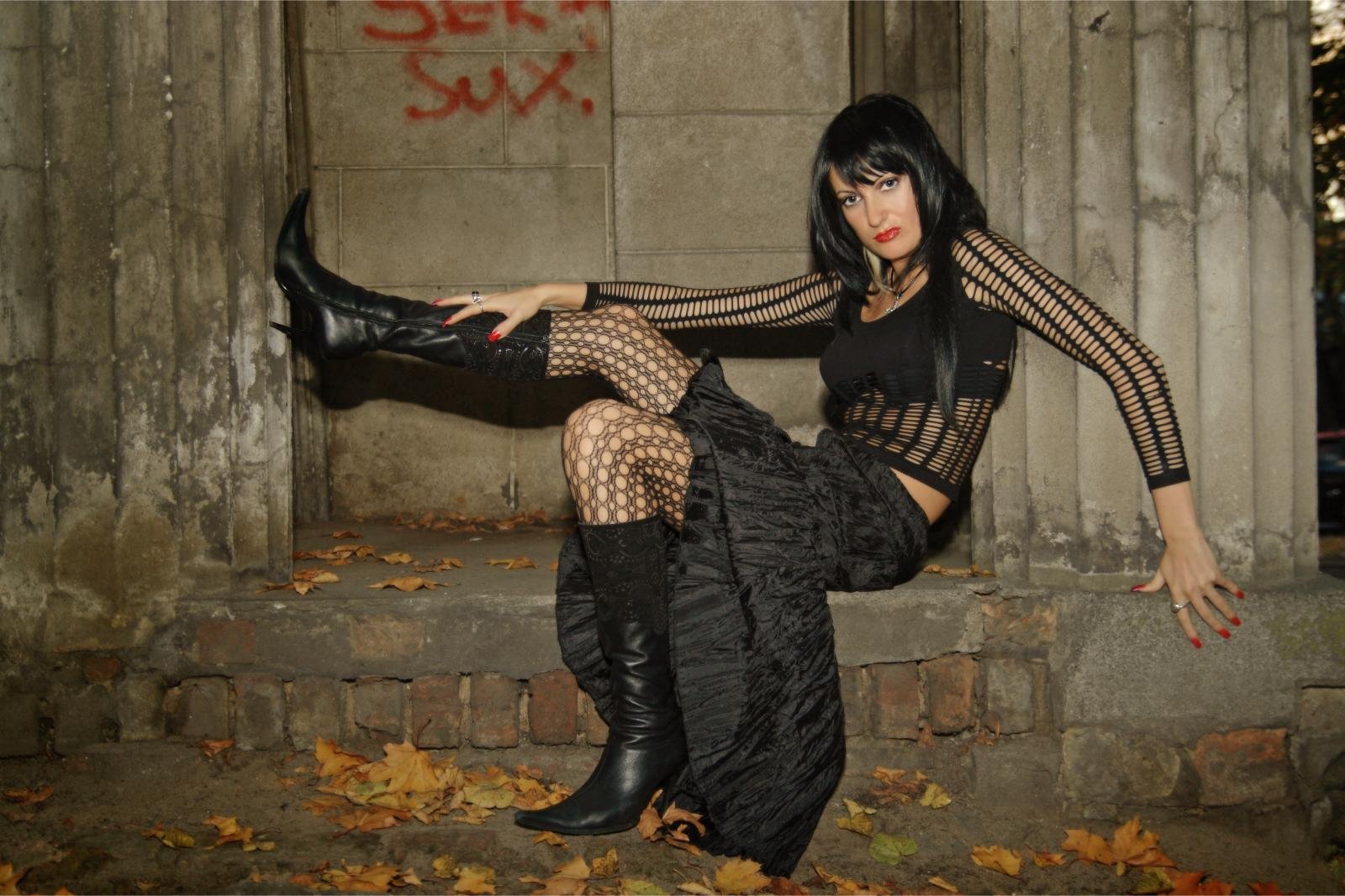 Goth woman wearing fishnet top, long black skirt, and boots sitting on stone steps outdoors.