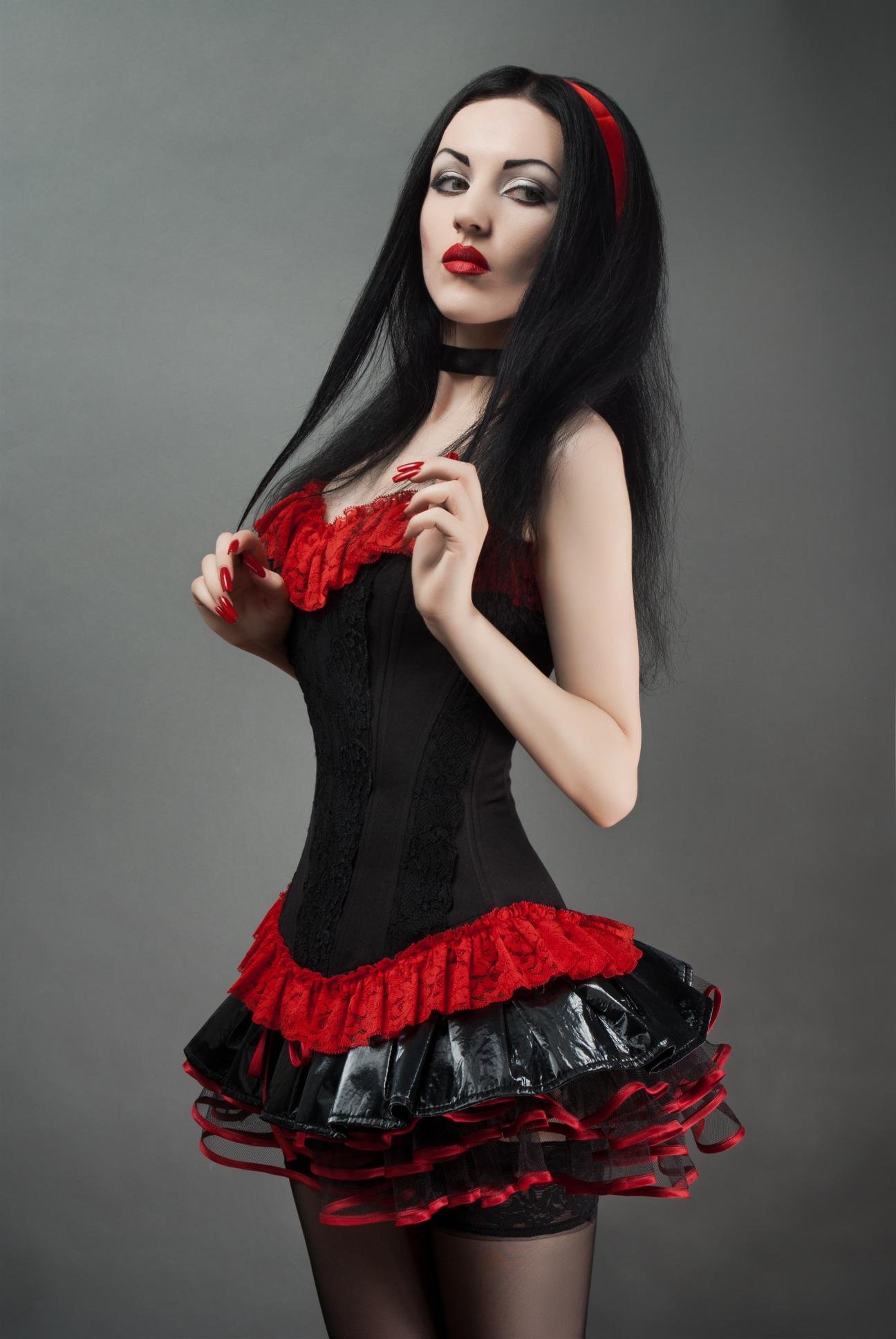 Gothic woman in red and black PVC skirt and corset posing confidently against grey background.
