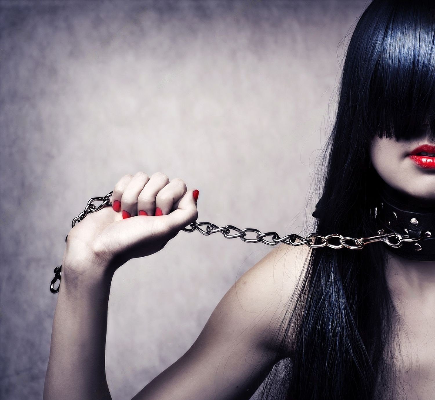 Woman with black hair holding metal chain and wearing collar, red lips and nails.